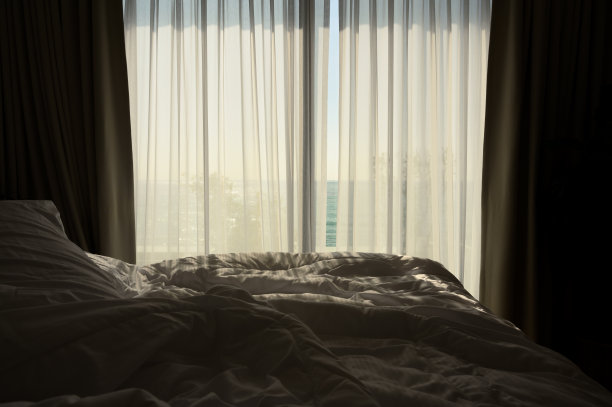 早上卧室靠窗。卧室里凌乱的床，白色的毯子和凌乱的枕头装饰。图片下载