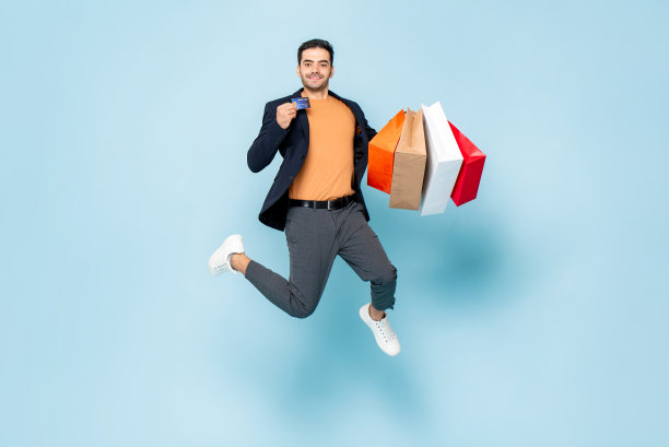 年轻微笑的白人男子的肖像持有购物袋和信用卡跳跃在孤立的淡蓝色工作室背景图片下载