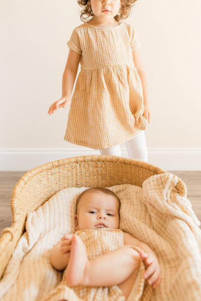 一个3岁的蹒跚学步的小女孩，穿着相配的黄色格子衣服，坐在一起，双臂举过5个月大的弟弟图片下载
