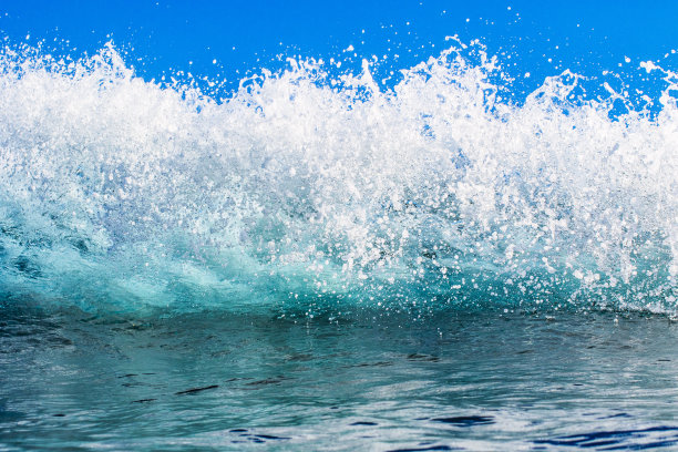 在晴朗的天空下，明亮的蓝色波浪创造泡沫白水图案的特写图片下载