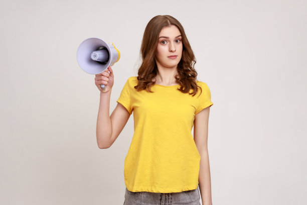 困惑困惑的十几岁的女孩棕色的头发在黄色的t恤手中拿着扩音器，找不到共同的语言，看着相机。图片下载