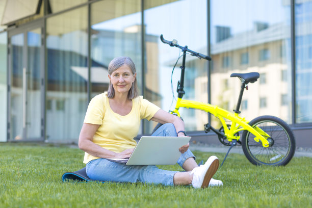教学。高级美女肖像坐在校园草地上与笔记本电脑和自行车。他学习和工作。他看着镜头，微笑着图片下载