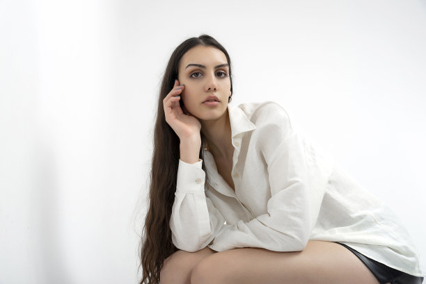 年轻美丽的照片模特穿着薄织物，夏天的白衬衫和白裤子，在白色背景的工作室里摆出艺术的姿势。图片下载