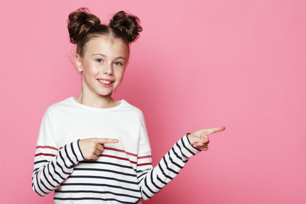 生活方式和童年概念:有趣的小女孩微笑着，在粉红色的背景上用手指指着右边。图片下载