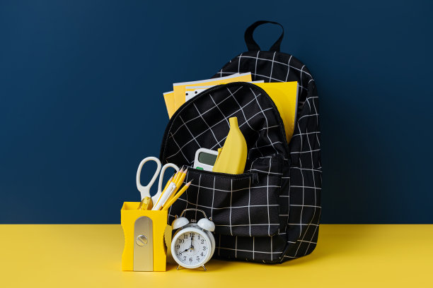 黑色格子双肩包与学校文具在黄色和蓝色的背景。办公用品和书包在桌子上。概念回到学校。图片下载