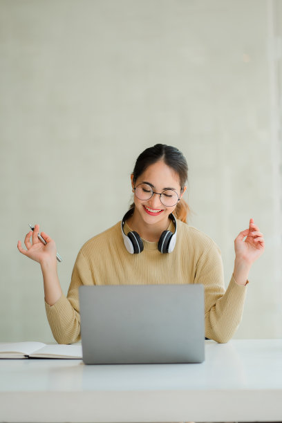 一个迷人和快乐的女亚洲大学生戴着耳机在她的脖子上坐在客厅的桌子微笑和工作在她的笔记本电脑。笔记本或自由职业商业想法垂直图片图片下载