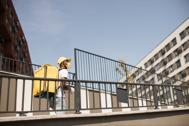 戴着口罩的帅气非洲裔美国快递员，戴着黄色帽子和背包的快递员图片下载