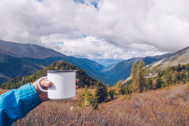 手握搪瓷白色马克杯模型与森林和山脉山谷背景。徒步用品和露营装备。Stock wildwood照片与白色金属杯。乡村产品模板。图片下载
