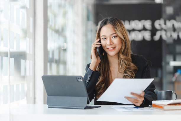 年轻的深色头发的亚洲女商人在用手机与客户交谈，看着相机，拿着工作表。图片下载
