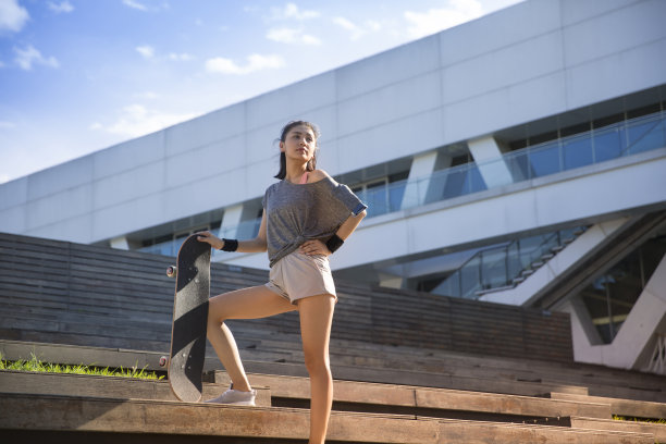 一个阳光明媚的夏日，一个年轻的女运动员拿着滑板站在城市建筑的楼梯上——照片图片下载
