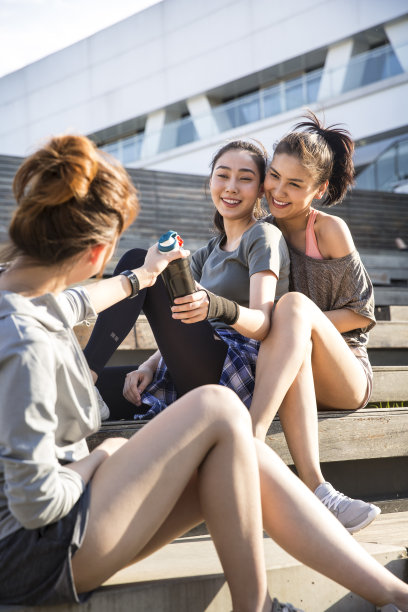 在一个阳光明媚的夏日，三位年轻的女士坐在城市里一座现代化建筑的台阶上，她们经过运动水瓶——照片图片下载