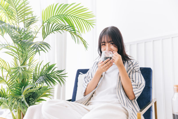 年轻漂亮的女人微笑着坐在现代生活区的椅子上，绿色的植物，放松地喝着一杯热咖啡，在一个舒适的家的室内环境中治愈倒影。图片下载