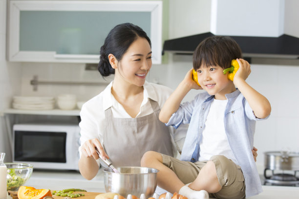 年轻的母亲在厨房里教儿子做饭，调皮的儿子把甜椒放在耳朵上当耳机。- - -库存图片图片下载