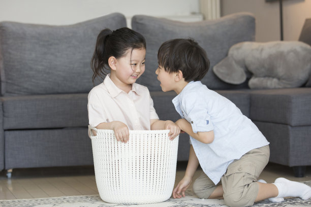 客厅的沙发旁，一个小女孩正准备坐在衣服筐里，小男孩和她开玩笑——库存照片图片下载
