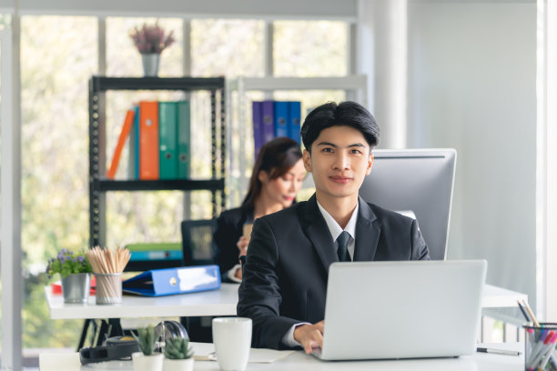 自信的商务人士坐在办公室与笔记本电脑工作，与团队在背景。图片下载