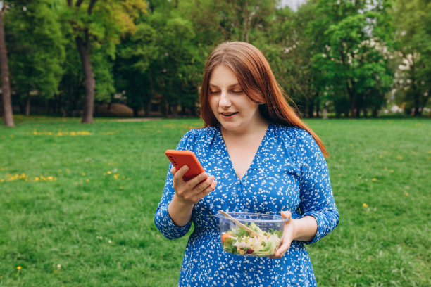 迷人的白人微笑女人吃沙拉的肖像。快乐年轻人用手机给客户服务手机app好评率。健康食品科技横幅图片下载