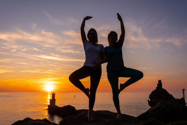 Vrksasana，两个女人在日落时的岩石上做冥想和瑜伽，旁边是海上的灯塔，健康和的生活图片下载