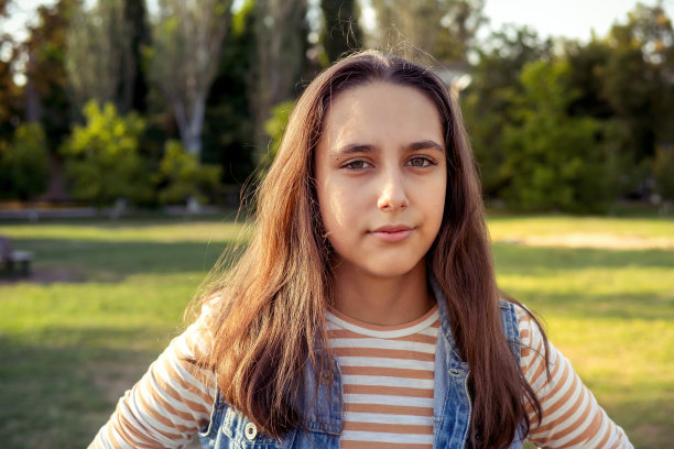 一个美丽开朗的十几岁学生女孩，棕色长发，穿着条纹长袖衬衫和牛仔背心在秋天的公园散步。自然照明黄金时间Z世代图片下载