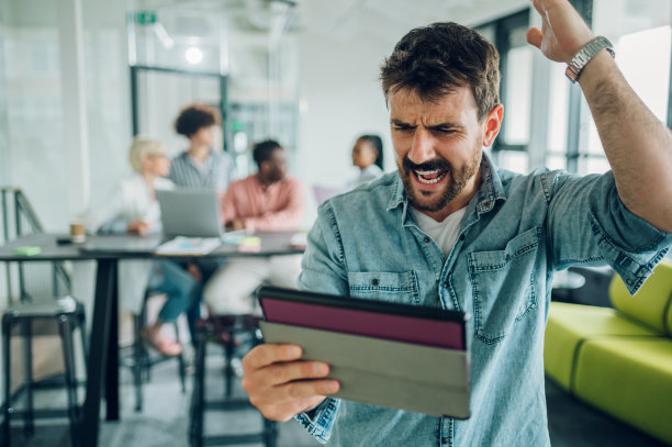 愤怒的商人在办公室里使用平板电脑，他的团队在后面工作图片下载