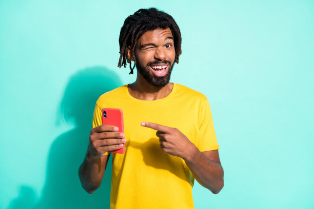 照片的年轻微笑非洲人与棕色脏辫眨眼的眼睛点手指手机孤立在蓝绿色的背景图片下载