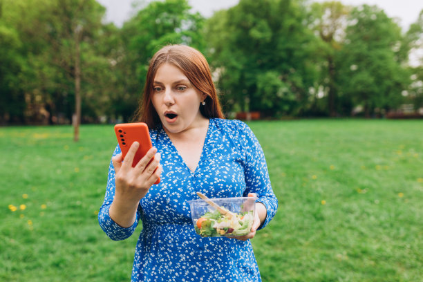迷人的白种人女人吃沙拉的画像。不开心的年轻人用手机给负率的客服手机app。健康食品和科技横幅图片下载