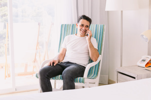年轻人坐在卧室的摇椅上，一边打电话一边微笑，光线透过窗户照进来。工作、度假、连接和智能手机的概念。图片下载
