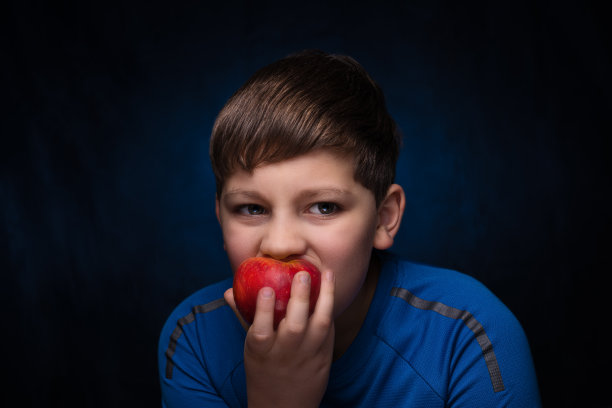 英俊的少年淡棕色头发在蓝色运动t恤吃多汁的红色苹果在蓝色孤立的背景图片下载