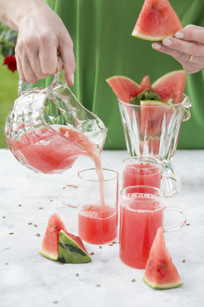 身穿绿色连衣裙的年轻女子从西瓜浆中倒出一杯红色清爽饮料，明亮自然的背景图片下载