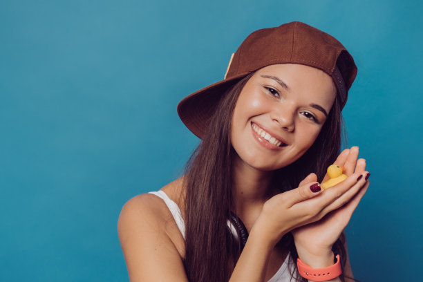 一个漂亮的女孩的特写肖像，长头发，戴着棒球帽，戴着耳机在她的脖子上，拿着一个小玩具鸭在蓝色的背景上拷贝空间。人的情绪。图片下载