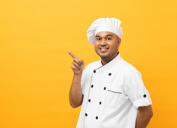 微笑年轻英俊的亚洲男子厨师在制服与帽子站立指着手指在黄色背景空白的文字。厨师职业厨师或面包师在厨房、餐厅和酒店工作的人。图片下载