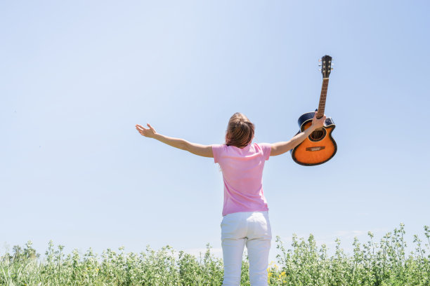 年轻女子站在草地上，手拿吉他，向天空举起手图片下载