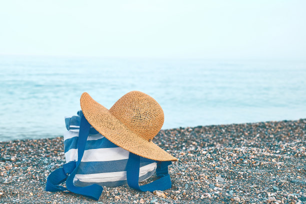 鹅卵石沙滩上的徒步者帆布蓝白条纹背包和草帽。旅行者放松度假的概念。图片下载