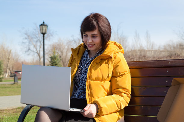 一位身穿亮橙色夹克的黑发成年女性坐在长椅上，呼吸着新鲜的空气，面带微笑地打开了一台新的笔记本电脑图片下载