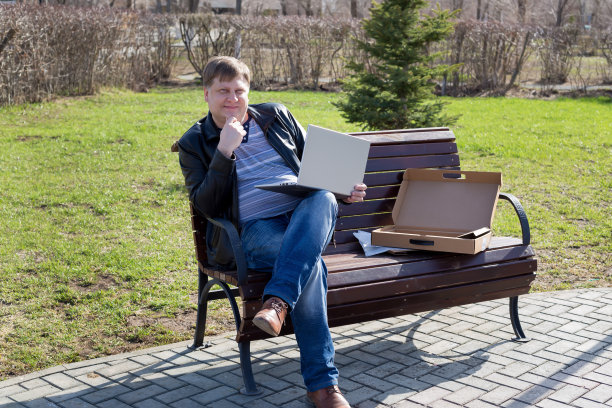 在一个阳光明媚的春日，穿着皮夹克的成年男性自由职业者坐在公园的长椅上，对着电脑工作图片下载