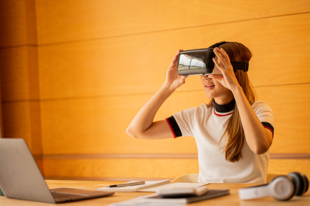 在家里戴着VR眼镜的开朗的白人女性。年轻女子享受眼镜与想象的手做想象的手势在虚拟现实设备在桌子上的笔记本电脑和计算器文件。图片下载