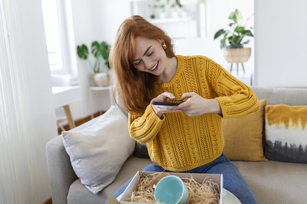 年轻满意的快乐女人购物狂客户坐在沙发上打开包裹快递箱，网购发货理念。拍摄产品照片发布在社交媒体上图片下载