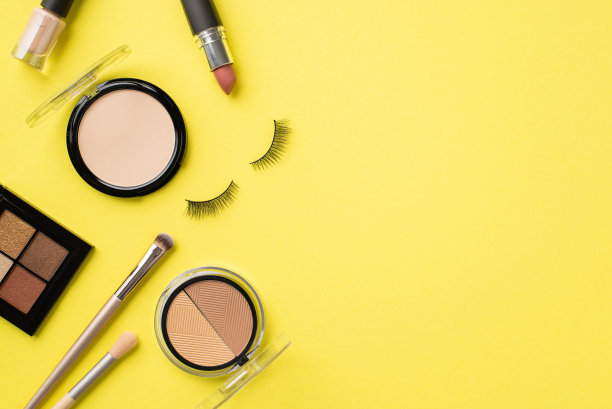 化妆的概念。俯视图照片紧凑的粉状眼影轮廓调色板指甲油化妆刷口红和假睫毛上孤立的黄色背景图片下载
