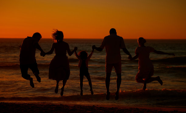 一个三代同堂的家庭在海滩上跳跃时的背影。一个快乐的家庭，有两个孩子，父母和祖父母牵着手在海滩上看日落图片下载