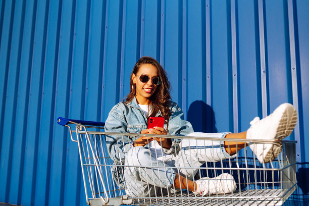年轻女子与电话坐在购物车有乐趣附近的彩色墙。图片下载