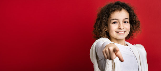 一个十岁的小白种人女孩与卷发前视图肖像特写站在红色背景前看着相机指着手指微笑幸福和快乐的复制空间图片下载