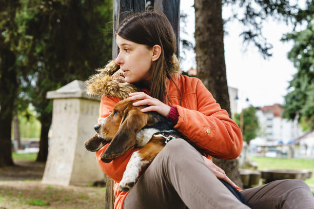 白人妇女抱着她的巴吉特猎犬宠物坐在公园里看着一边，在冬天或春天穿着夹克真实的人复制空间图片下载
