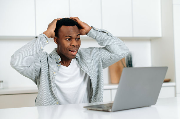 惊喜的年轻成年商人非洲裔美国人，经理，经纪人以惊讶的姿势坐在桌子旁，使用笔记本电脑，被好消息或消息震惊，得到了经济利润，兴奋。图片下载