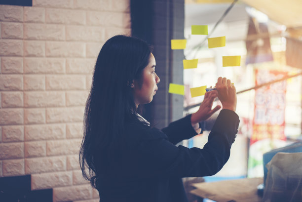 亚洲女商人把创意和计划写在玻璃墙的便签纸上。项目战略计划将目标任务和创意写在便利贴上，并将便条贴在办公室的玻璃板上图片下载