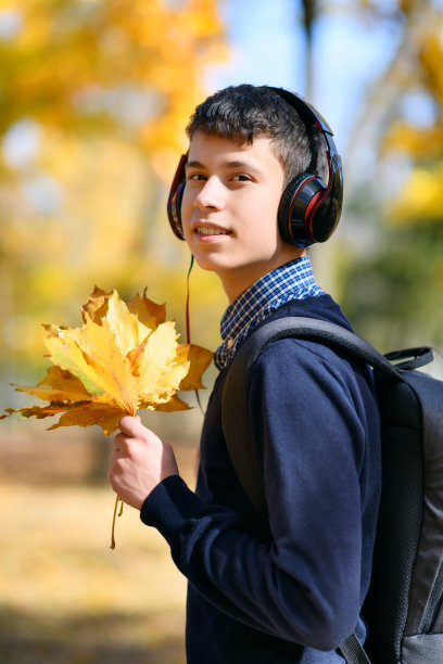 一个十几岁的男孩走在秋天的公园里，戴着耳机听着音乐，阳光明媚的日子里，树上的叶子都泛黄了图片下载
