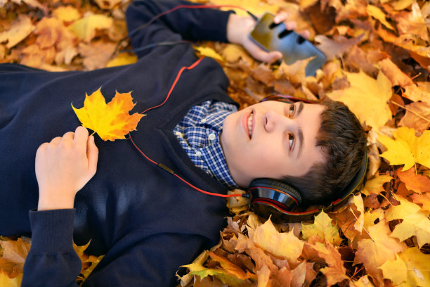 一个十几岁的男孩戴着耳机听着音乐，躺在黄色的秋天的枫叶上，一个阳光明媚的日子，美丽的秋天的自然图片下载
