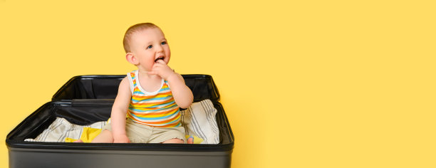 婴儿蹒跚学步的男孩坐在一个手提箱里，衣服，工作室黄色的背景。一个孩子在为度假旅行打包行李时，在一个大袋子里玩耍，复印空间。一岁(12个月)图片下载