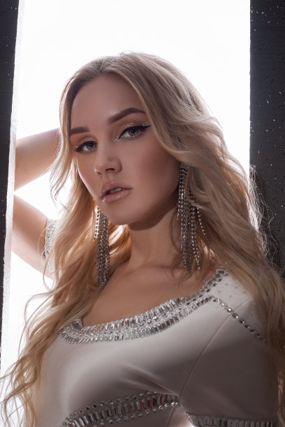 美丽的美女金发卷曲的长发女子在一件紧身的白色短连衣裙的背景下打开在黑色的墙壁。长长的金发在风中飘动图片下载