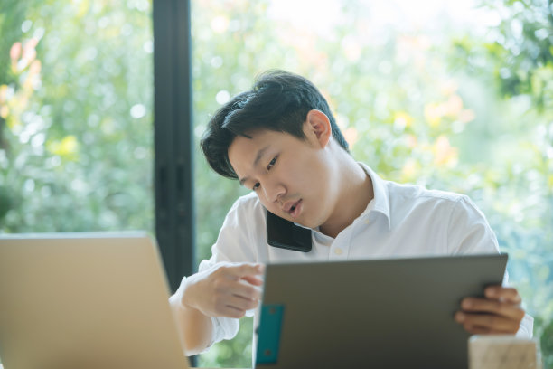 一名亚洲男子一边忙着工作，一边在家打电话和使用笔记本电脑图片下载