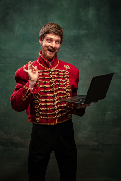 英俊的年轻人在中世纪骠骑兵的形象使用笔记本电脑隔离在深蓝色的背景。复古的风格，比较时代的概念。图片下载