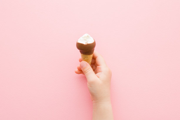 女婴手握华夫饼筒白色香草冰淇淋与黑巧克力釉在浅粉红色的桌子背景。柔和的颜色。特写镜头。冷甜的零食。咬的食物。自顶向下的观点。图片下载